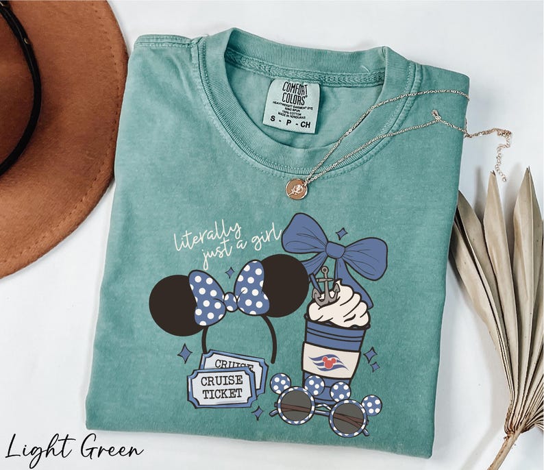A light green t-shirt with a graphic design of a cruise ticket, a pair of sunglasses, and a cup with a bow, along with a necklace and a hat.