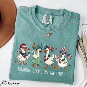 A light green t-shirt with a patriotic goose design and the text "Patriotic Goose on the Loose" printed on it, along with a necklace and a brown hat.