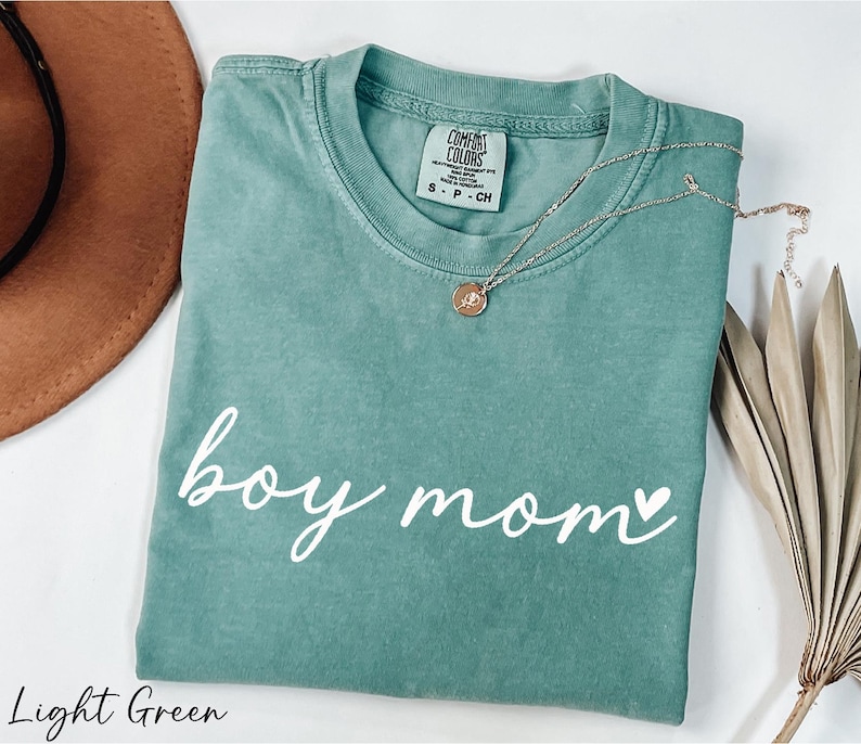 A light green t-shirt with the text "boy mom" printed on it, along with a necklace and a brown hat, all placed on a white surface.