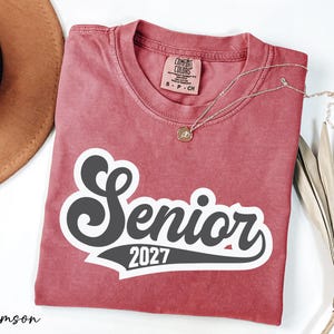 a red t-shirt with the text "SENIOR 2027" printed on it, along with a necklace and a hat.