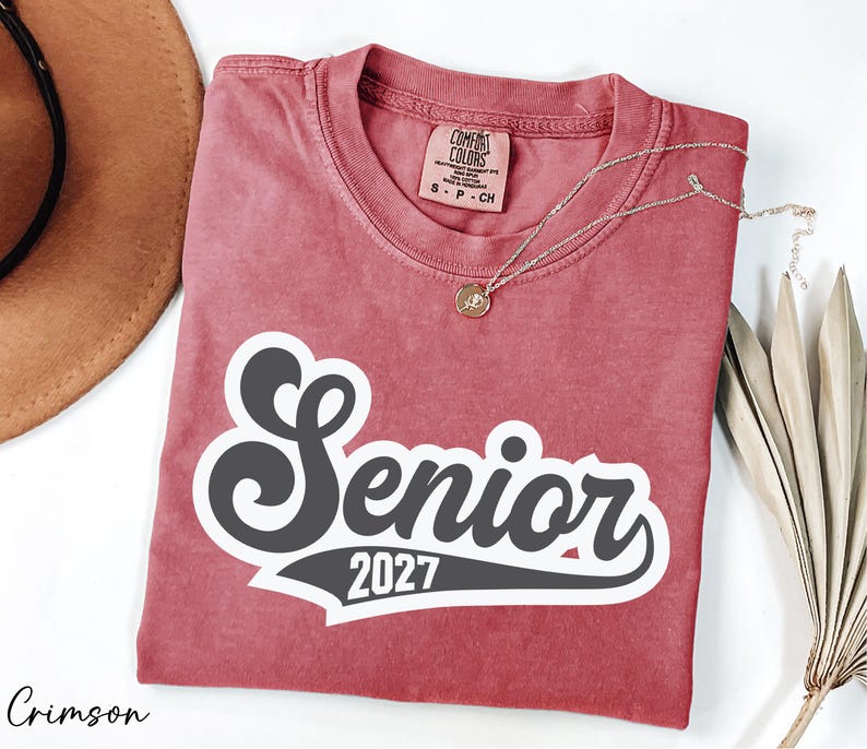 a red t-shirt with the text "SENIOR 2027" printed on it, along with a necklace and a hat.