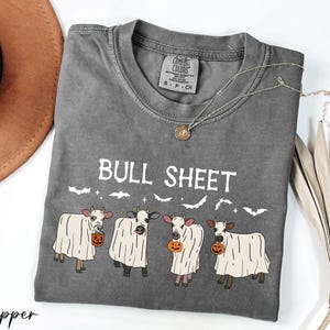 a gray t-shirt with a Halloween-themed design, including a group of cows dressed in ghost costumes and holding pumpkins. The t-shirt is accompanied by a brown hat, a necklace, and some dried leaves, creating a festive and spooky atmosphere.