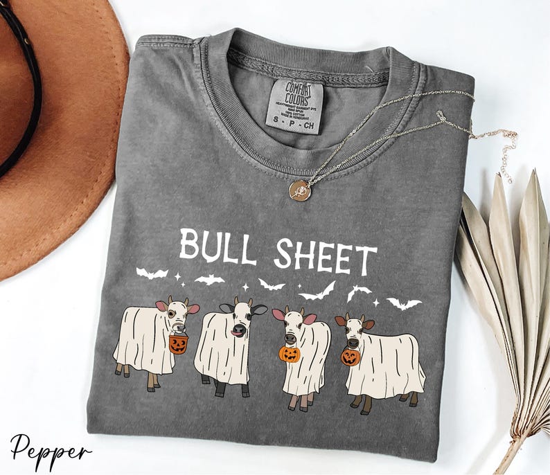 a gray t-shirt with a Halloween-themed design, including a group of cows dressed in ghost costumes and holding pumpkins. The t-shirt is accompanied by a brown hat, a necklace, and some dried leaves, creating a festive and spooky atmosphere.