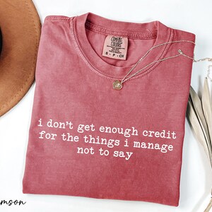 a red t-shirt with white text that reads "I don't get enough credit for the things I manage not to say", accompanied by a brown hat and some dried leaves.