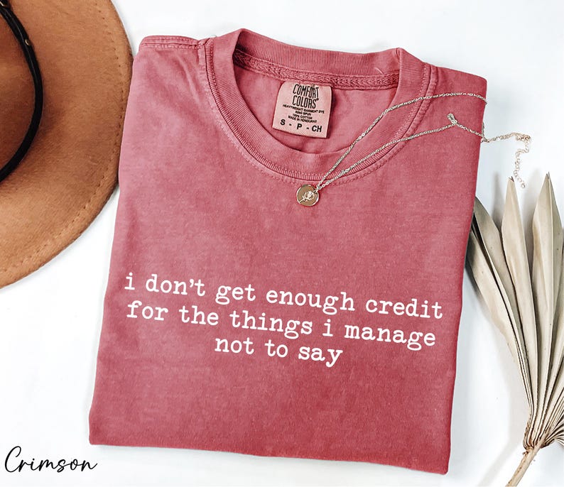 a red t-shirt with white text that reads "I don't get enough credit for the things I manage not to say", accompanied by a brown hat and some dried leaves.