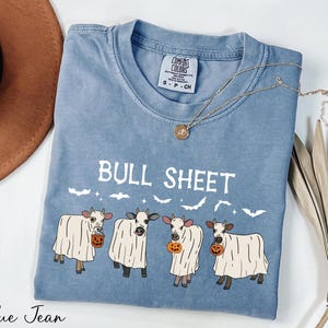 A blue t-shirt with a graphic design of four cows dressed in ghost costumes, along with the text "Bull Sheet" and a pumpkin. The t-shirt is placed on a white surface, with a brown hat and some dried leaves nearby.