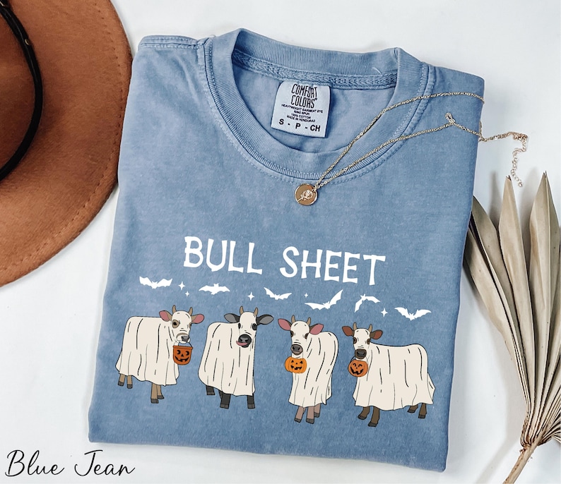 A blue t-shirt with a graphic design of four cows dressed in ghost costumes, along with the text "Bull Sheet" and a pumpkin. The t-shirt is placed on a white surface, with a brown hat and some dried leaves nearby.