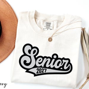 a white t-shirt with a black and white graphic design, a brown hat, a necklace, and some dried leaves, all arranged on a white background.