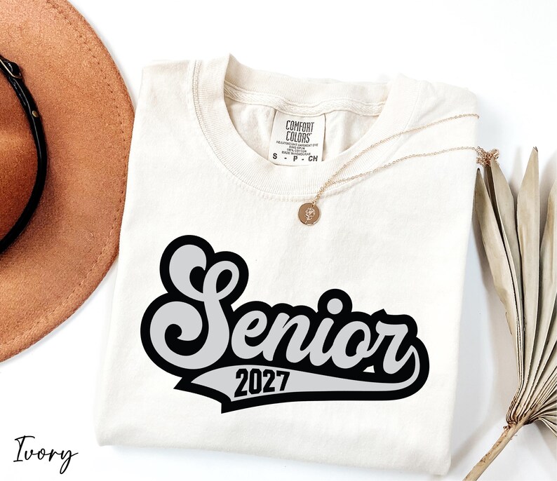 a white t-shirt with a black and white graphic design, a brown hat, a necklace, and some dried leaves, all arranged on a white background.