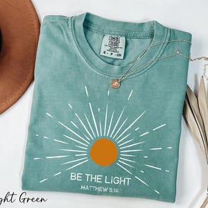 K&ouml;nnte beinhalten: Hellgr&uuml;nes T-Shirt mit einem wei&szlig;en Sonnenstrahlen-Design und dem Text "Sei das Licht Matth&auml;us 5:16".