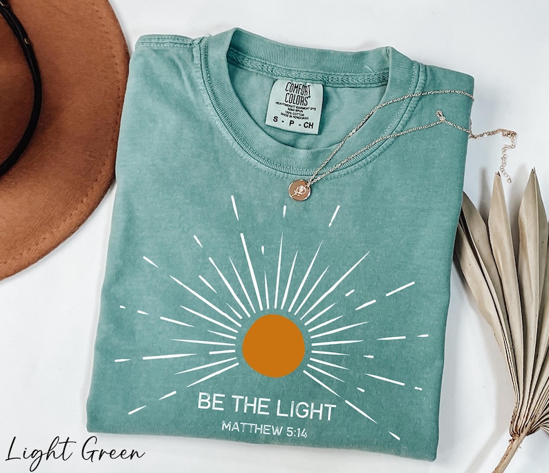 K&ouml;nnte beinhalten: Hellgr&uuml;nes T-Shirt mit einem wei&szlig;en Sonnenstrahlen-Design und dem Text "Sei das Licht Matth&auml;us 5:16".