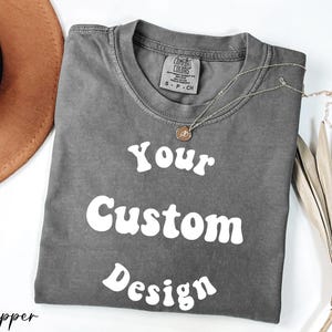 Op de afbeelding: Een grijs T-shirt met de tekst "Your Custom Design" in wit, retro-stijl lettertype. Het shirt is gemaakt van een zacht, comfortabel materiaal.