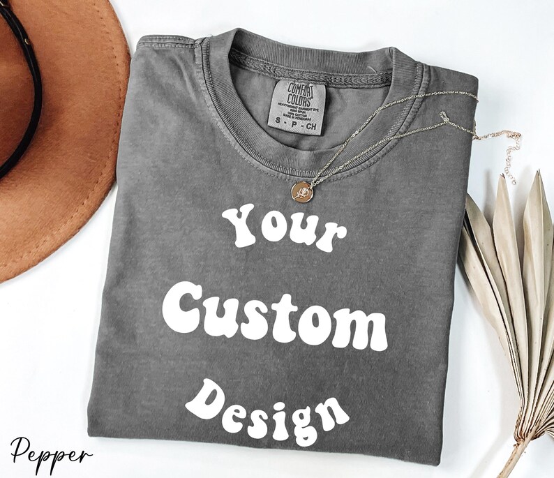 Op de afbeelding: Een grijs T-shirt met de tekst "Your Custom Design" in wit, retro-stijl lettertype. Het shirt is gemaakt van een zacht, comfortabel materiaal.
