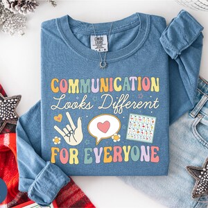May include: Blue long-sleeve shirt with the text "COMMUNICATION Looks Different FOR EVERYONE" in a colourful retro font. The shirt features a hand in sign language, a heart, and a communication chart. A silver necklace is visible.