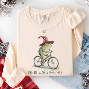 May include: Ivory long-sleeve shirt with a whimsical illustration of a green frog wearing a red wizard hat and riding a bicycle. The text "OFF TO CAUSE A KERFUFFLE" is printed below the frog. The shirt is folded and displayed with denim jeans and decorative stars.