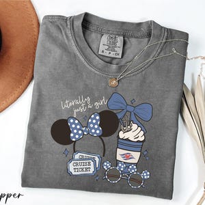 A gray t-shirt with a graphic design featuring a cup with a blue ribbon, sunglasses, and a pair of Mickey Mouse ears, along with the text "Literally just a girl" and "Cruise Ticket".