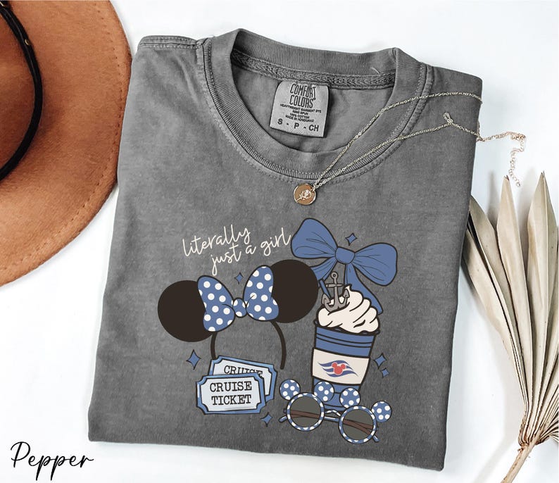 A gray t-shirt with a graphic design featuring a cup with a blue ribbon, sunglasses, and a pair of Mickey Mouse ears, along with the text "Literally just a girl" and "Cruise Ticket".