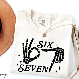A white t-shirt with a graphic design of a skeleton hand and the text "Six Seven" printed on it, along with a gold necklace and a brown hat.