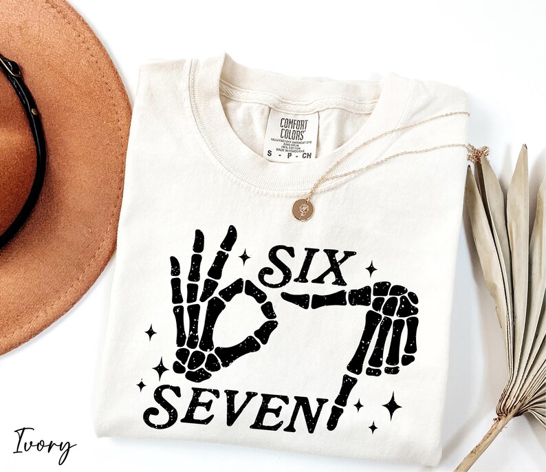 A white t-shirt with a graphic design of a skeleton hand and the text "Six Seven" printed on it, along with a gold necklace and a brown hat.