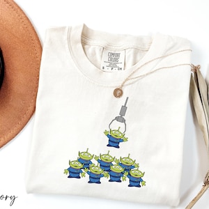 May include: A white t-shirt with a graphic of the green alien toys from Toy Story. The aliens are being lifted by a claw machine. The shirt has a tag that says "Comfort Colors" and "S - P - CM".
