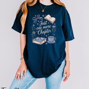 A woman wearing a navy blue t-shirt with a graphic design that says "Just one more Chapter" and includes illustrations of a book, a cup of coffee, and flowers.