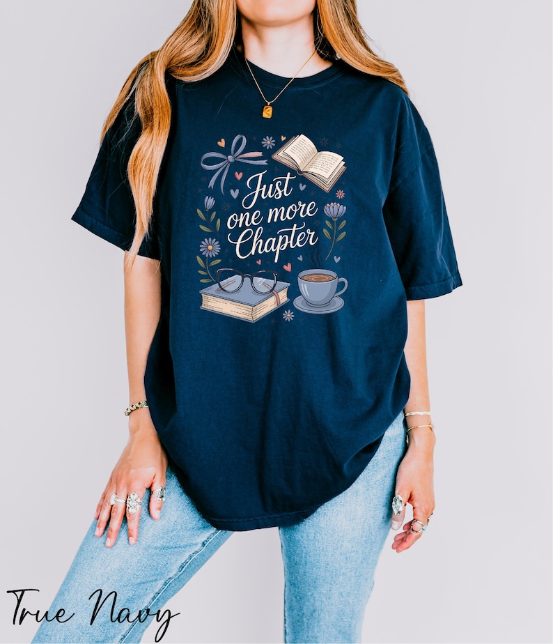 A woman wearing a navy blue t-shirt with a graphic design that says "Just one more Chapter" and includes illustrations of a book, a cup of coffee, and flowers.