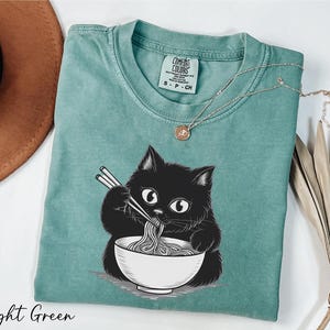 A light green t-shirt with a black cat illustration and a bowl of noodles, accompanied by a brown hat and some dried leaves.