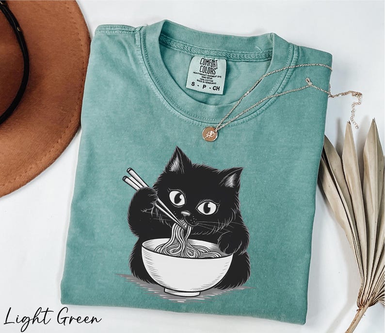 A light green t-shirt with a black cat illustration and a bowl of noodles, accompanied by a brown hat and some dried leaves.