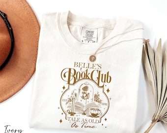 May include: Ivory t-shirt with a gold graphic design that says "Belle's Book Club" with a rose, book, and teacup. The text also says "Est. 1991" and "Tale as old as time."