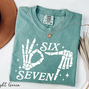 A light green t-shirt with a skeleton hand print and the text "Six Seven" printed on it, along with a necklace and a brown hat.
