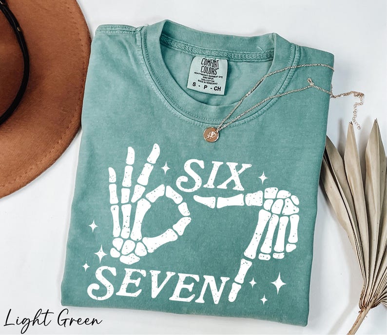 A light green t-shirt with a skeleton hand print and the text "Six Seven" printed on it, along with a necklace and a brown hat.
