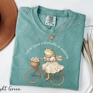 A light green t-shirt with a printed image of a frog riding a bicycle, along with a necklace and a hat.