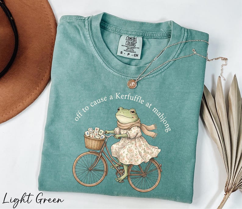 A light green t-shirt with a printed image of a frog riding a bicycle, along with a necklace and a hat.