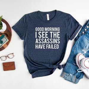 May include: Navy blue t-shirt with the text "GOOD MORNING I SEE THE ASSASSINS HAVE FAILED" in white. The shirt is paired with blue jeans, white sneakers, and accessories.