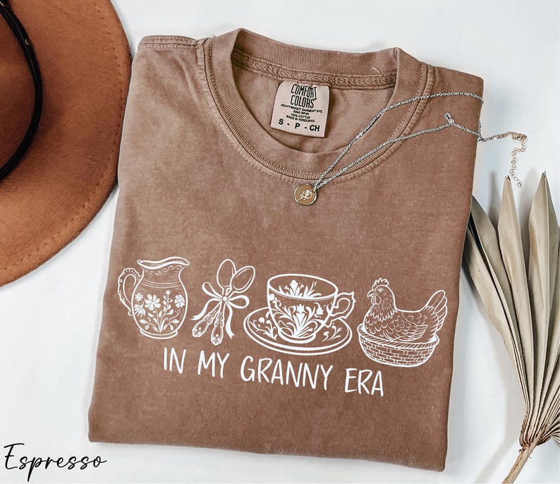 A brown t-shirt with a printed design of various kitchen items, including a teapot, a cup, a bowl, and a spoon, along with the text "In my Granny Era" printed on it. The t-shirt is laid out on a white surface, and there is a hat and some dried leaves nearby.