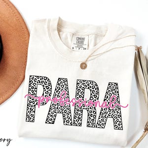 a white t-shirt with the word "PARA" printed in pink and black leopard print letters, along with the word "professional" written in pink cursive below it. The t-shirt is placed on a white surface, and there is a brown hat and some dried leaves nearby.