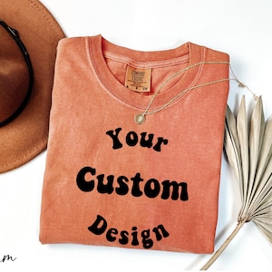 Op de afbeelding: Een koraalkleurig T-shirt met de tekst "Your Custom Design" in zwart retro-lettertype.