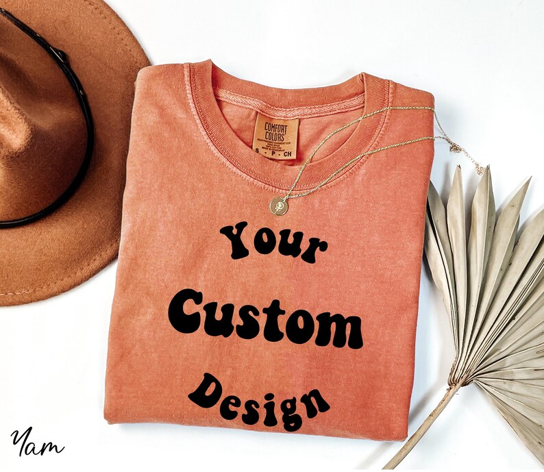 Op de afbeelding: Een koraalkleurig T-shirt met de tekst "Your Custom Design" in zwart retro-lettertype.