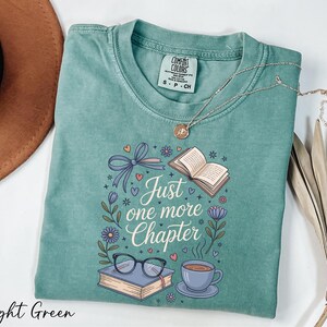 A light green t-shirt with a printed design, a pair of glasses, a book, a cup of coffee, and a plant, along with a necklace and a brown hat.