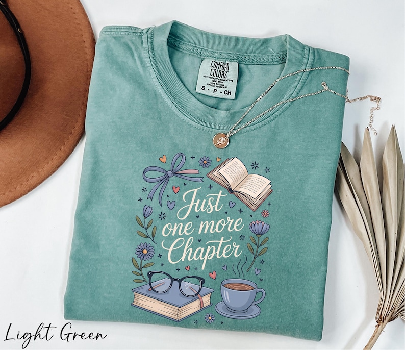 A light green t-shirt with a printed design, a pair of glasses, a book, a cup of coffee, and a plant, along with a necklace and a brown hat.