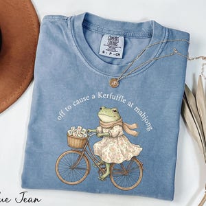 A blue t-shirt with a cartoon image of a frog riding a bicycle, along with a necklace and a hat.