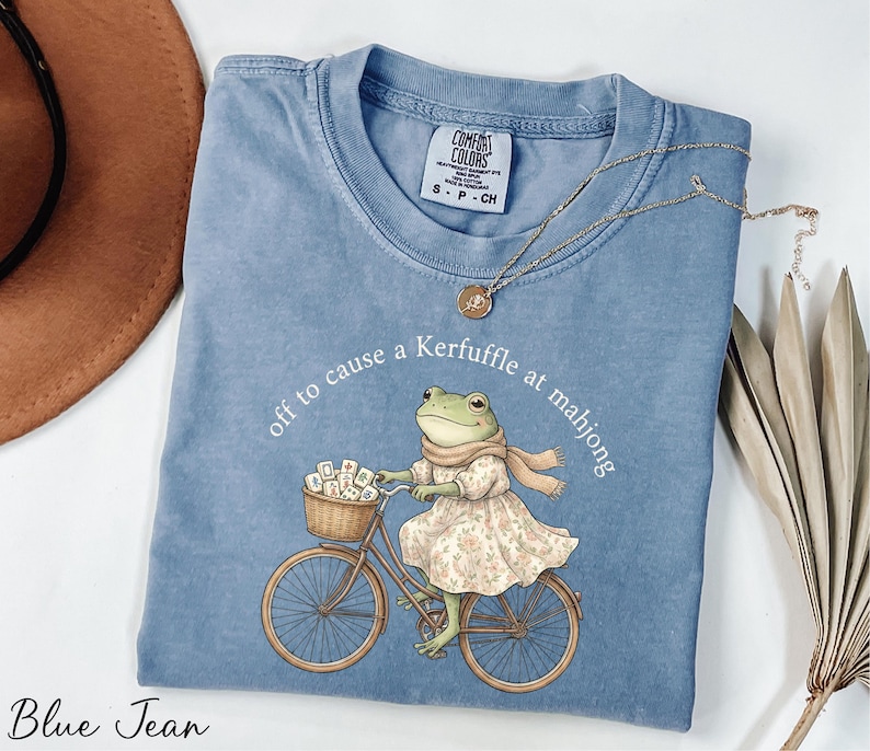 A blue t-shirt with a cartoon image of a frog riding a bicycle, along with a necklace and a hat.
