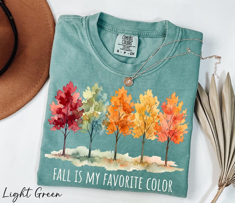 A light green t-shirt with a colorful autumn tree design and the text "FALL IS MY FAVORITE COLOR" printed on it, along with a necklace and a brown hat.