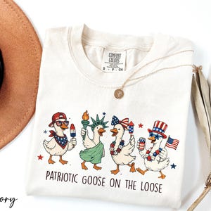 A white t-shirt with a patriotic goose design and the text "Patriotic Goose on the Loose" printed on it, along with a necklace and a hat.