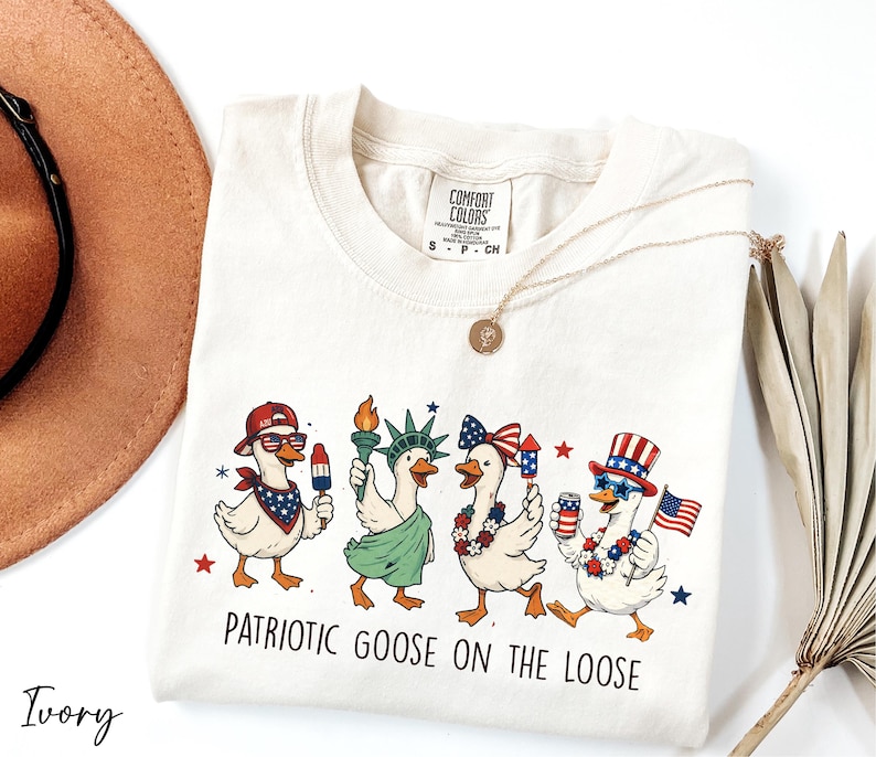 A white t-shirt with a patriotic goose design and the text "Patriotic Goose on the Loose" printed on it, along with a necklace and a hat.