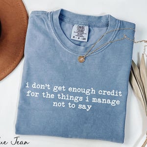 a blue t-shirt with white text that reads "I don't get enough credit for the things I manage not to say". The t-shirt is laid out on a white surface, accompanied by a brown hat and some dried leaves.
