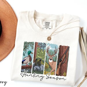 A white t-shirt with a graphic design depicting a deer, a bird, and a tree, along with the text "Hunting Season".