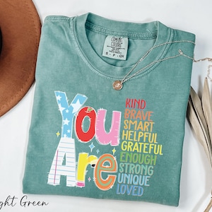 May include: Light green t-shirt with the words "You Are" in colorful block letters and a list of positive adjectives. A gold necklace with a pendant rests on the shirt. The shirt is labeled "Comfort Colors".
