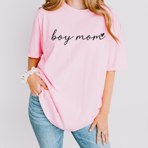 A woman wearing a pink t-shirt with the text "boy mom" printed on it, standing against a white background.