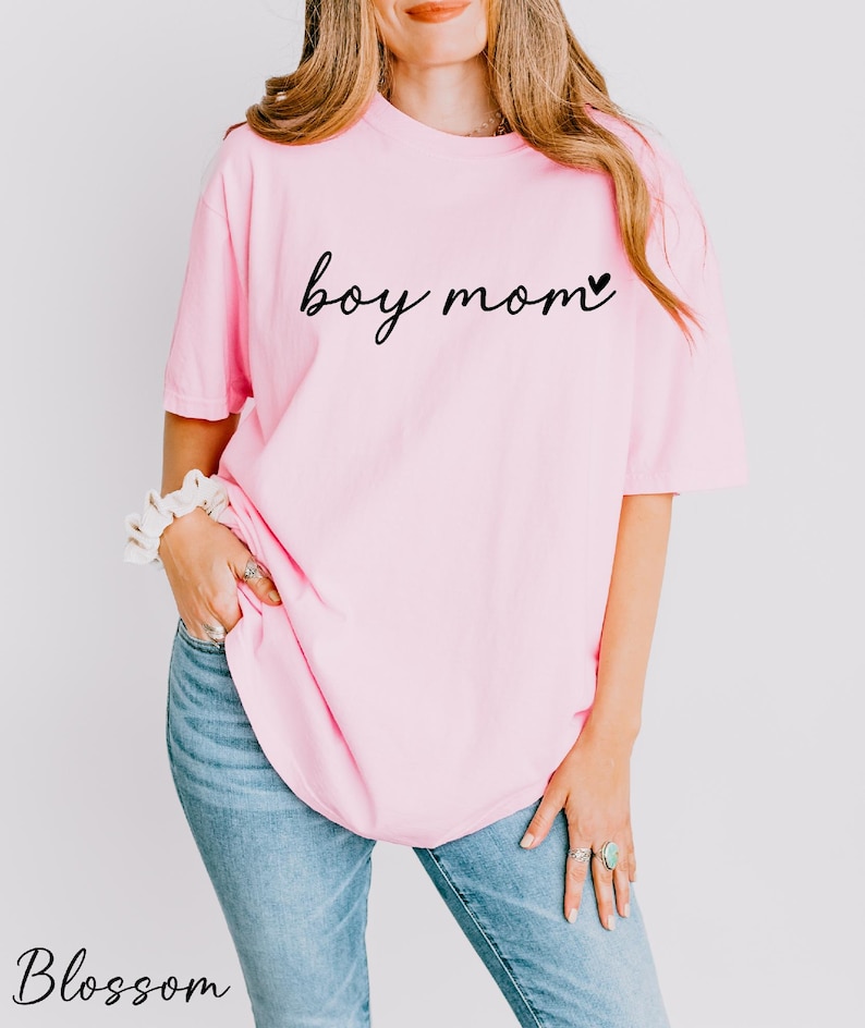 A woman wearing a pink t-shirt with the text "boy mom" printed on it, standing against a white background.